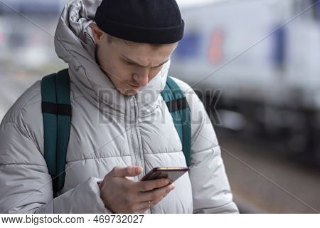 Young Man Using Smartphone, Texting, Cheks The Route Of Trip On Google Mapes While Waiting Train At 