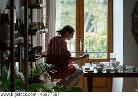 Thoughtful Creative Woman Artist Decorating Clay Plates After Kiln Firing Sits At Table In Workshop.