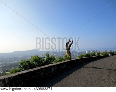 A Man Wearing A Hat, T-shirt, Pants, And Slippers Is Performing A Handstand At The Tantalus Mountain
