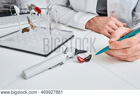 Audioprosthetist Presentation Hearing Aids In Two Colors To His Patient Lying On Table Near Otoscope