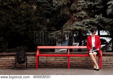 Attractive Businesswoman Is Doing Notes In Paper Notebook On Bench In The Park. Executive In Red Bla