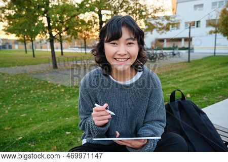 Young Asian Girl With Graphic Pencil And Tablet, Sits In Park On Bench, Draws Scatches, Does Her Hom