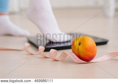 Woman's Foot In White Yjcrt Stepping On A Scale With A Measuring Tape And Fresh Peach Fruit. The Con