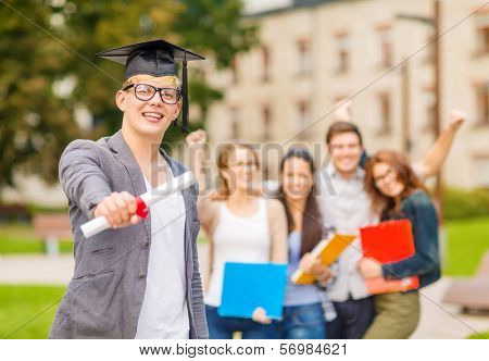 education, campus and teenage concept - smiling teenage boy in corner-cap and eyeglasses with diploma and classmates on the back