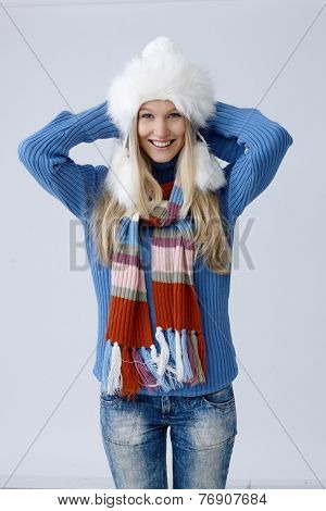 Happy young blonde woman standing in hat and scarf, folding arms around scruff, smiling, looking at camera.