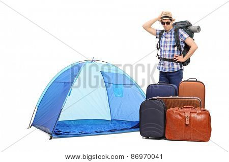 Baffled male tourist standing next to a tent and five huge briefcases isolated on white background