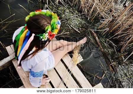 beautiful young girl in a white chemise and a wreath of flowers on her head sitting on a wooden bridge on a background of the river. The Celebration Of Midsummer