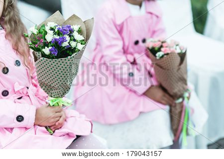 Little girls in pink coats sit on the white chairs holding bouquets in their arms