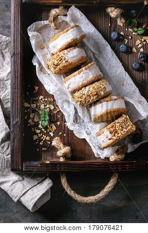Set of homemade ice cream sandwiches in oat cookies with almond sugar crumbs on baking paper in woden tray over dark metal texture background. Top view with space