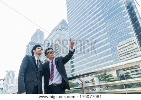 Two young businessmen talking and pointing at something while standing within the city.