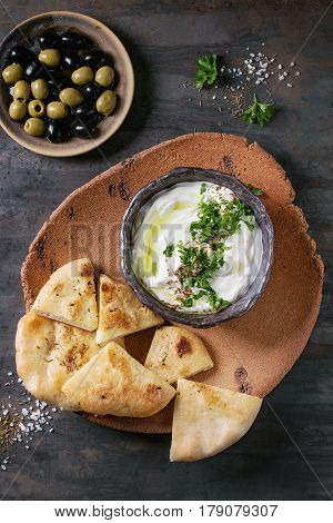 labneh middle eastern lebanese cream cheese dip with olive oil, salt, herbs served with olives, traditional pita bread on terracotta plate over dark texture metal background. Top view with space