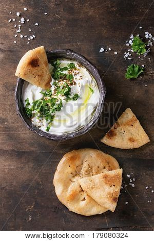 labneh middle eastern lebanese cream cheese dip with olive oil, salt, herbs served with traditional pita bread in terracotta bowl over dark texture wooden background. Top view with space