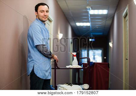 A man who is on the hotel cleaning crew staff is smiling with a towel, pillow and vacuum in the process of cleaning the hotel rooms and delivering top-knotch service to the guests