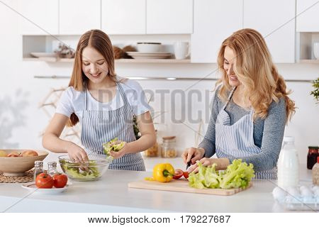 Healthy receipts. Pleasant positive teenage girl and her mother standing in the kitchen and cooking salad while eating healthy food