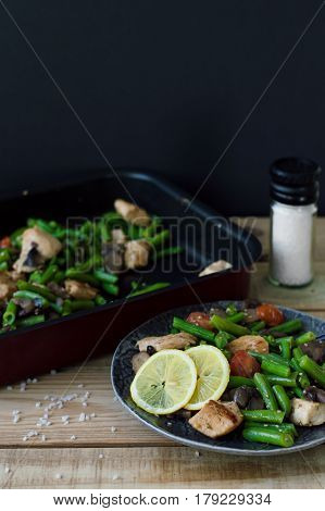Vertical photo of hot salad with green beans chicken breasts mushrooms cherry tomatoes and sesame served with two lemon slices on grey plate