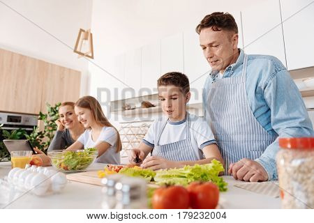 Involved in culinary. Pleasant little boy cooking with his father while mother and daughter sitting at th etable and using tablet