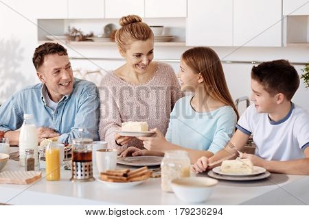 Help yourself. Cheerful loving mother serving breakfast to her family members while sitting in the kitchen together