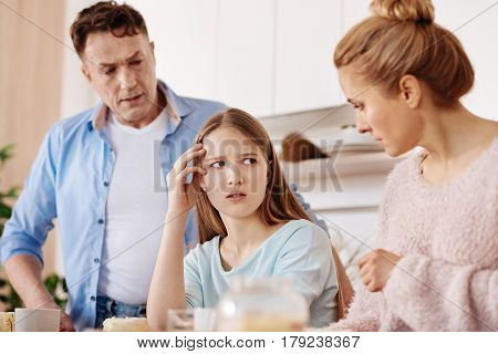 Need to solve problem. Gloomy thoughtful girl sitting at the table and discussing her problems with her parents in the kitchen