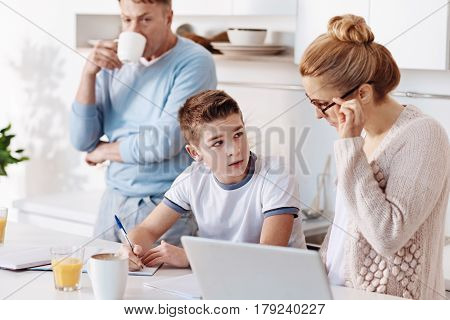 Control everything. Diligent boy doing home assignment while her mother controlling him while sitting in the kitchen together