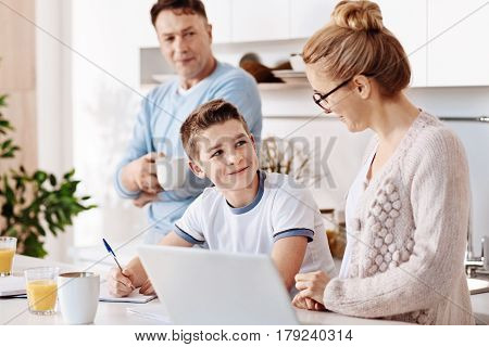 Good result. Positive diligent boy sitting at the table and doing his home work while his mother feeling ptoud of him