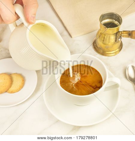 A square photo of milk being poured into a cup of coffee, with vintage coffee grinder and pot in the background, and cane sugar. Selective focus, toned image