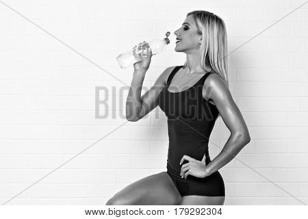 Portrait of blonde woman in black bodysuit with bottle of water posing with dumbbells.