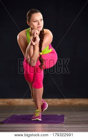 Beautiful young woman working out indoors, doing yoga exercise on violet mat, standing in Vrksasana Posture, Tree Pose. Woman in the sportwear, pink leggings and green t-shirt. Healthy lifestyle.