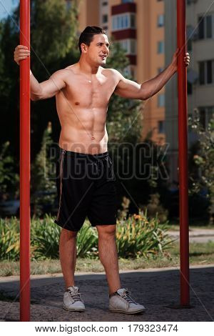 Young strong athlete standing at horizontal bar against buildings. Sportsman in black short and white sneakers. Healthy lifestyle.
