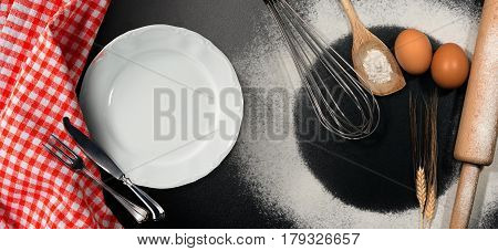 Baking background with empty plate and silver cutlery on a black table with rolling pin whisk eggs spoons ears of wheat and a checkered tablecloth