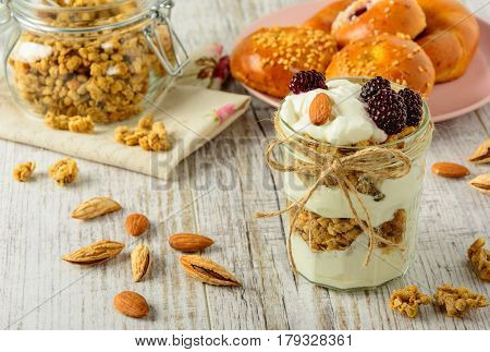 Healthy breakfast ingredients. Homemade granola with yoghurt in open glass jar and fresh fragrant buns. The food is rich in useful carbohydrates vitamins and nutrients that are essential for health. The dishes stand on a white wooden table.