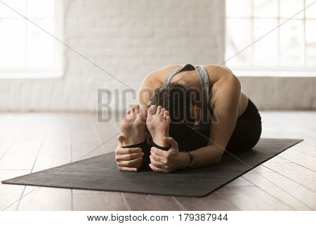 Young attractive yogi woman practicing yoga concept, sitting in paschimottanasana exercise, Seated forward bend pose, working out on black mat, wearing sportswear, white loft studio background