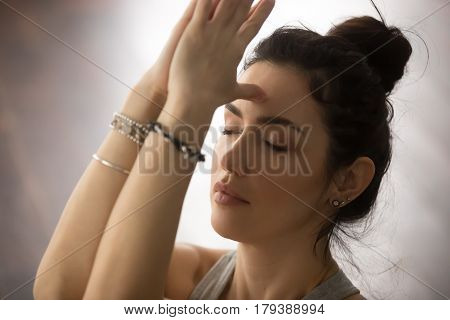 Young attractive yogi woman practicing yoga concept, doing namaste gesture, namaste hands to forehead with her eyes closed, working out, wearing wrist bracelets, studio background, closeup portrait