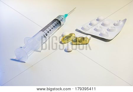 A variety of medication including a syringe with a green tip blister pack white pills and yellow plastic pills full of cod liver oil vitamin D supplement shot at a low angle against a white background