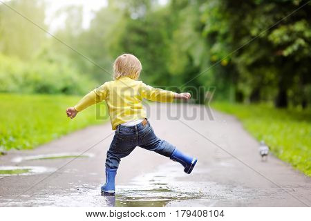 Little Boy Wearing Rain Boots Jumping In Pool Of Water