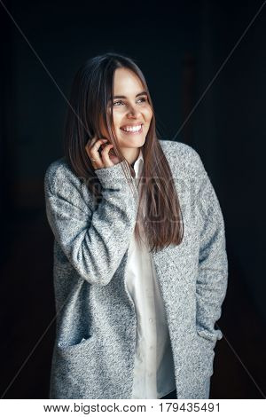 Portrait of smiling white Caucasian brunette young beautiful girl woman model with dark hair and brown eyes in white shirt and grey long coat looking in camera on black background