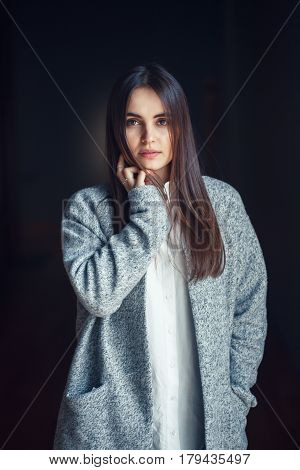 Portrait of pensive white Caucasian brunette young beautiful girl woman model with dark hair and brown eyes in white shirt and grey long coat looking in camera on black background