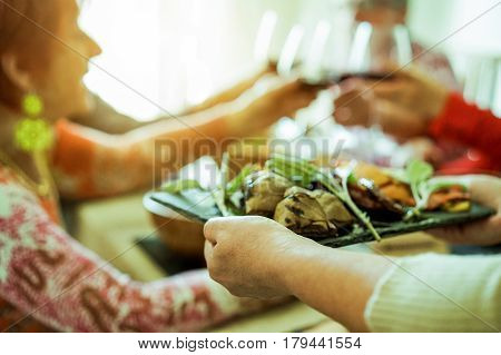 Chef woman serving dry tomatoes, cooked artichokes,peppers with old people cheering in background - Happy senior people having lunch toasting red wine - Focus on left dish hand - Warm vintage filter