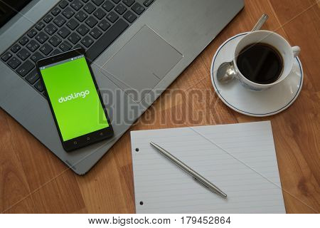 Nitra, Slovakia, march 28, 2017: Duolingo application in a mobile phone screen. Workplace with a laptop, an earphones, notepad, pen and coffee on wooden background
