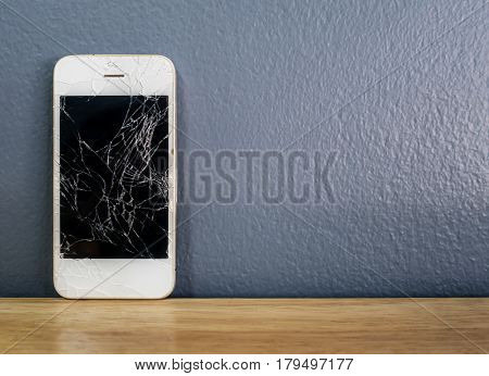 Front view image of white broken cracked screen smartphone leaning against the gray wall on wooden table with copy space