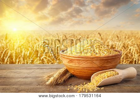 Bulgur or couscous in ceramic bowl and ears of wheat on table with ripe cereal field on the background. Agriculture and harvest concept. Golden wheat field on sunset