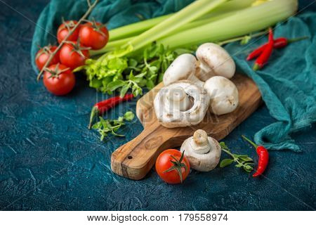 Fresh mushrooms champignons on a wooden cutting board parsley greens celery stalks cherry tomatoes and hot red chili pepper on a dark blue background