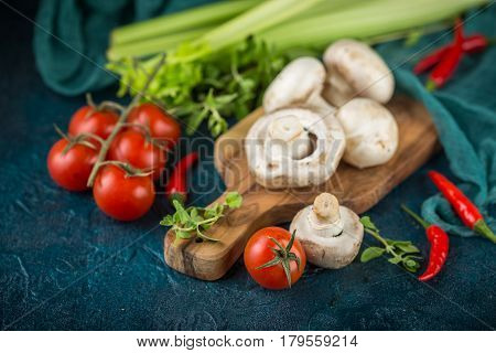 Fresh mushrooms champignons on a wooden cutting board parsley greens celery stalks cherry tomatoes and hot red chili pepper on a dark blue background