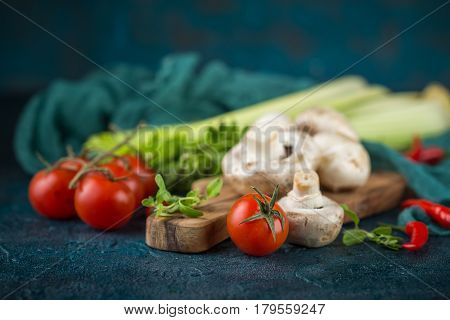 Fresh mushrooms champignons on a wooden cutting board parsley greens celery stalks cherry tomatoes and hot red chili pepper on a dark blue background
