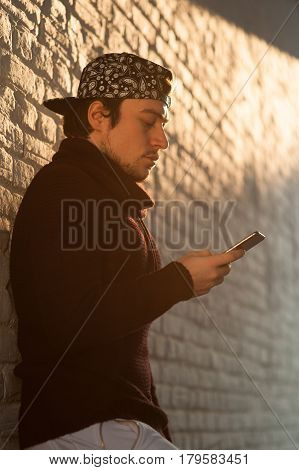 young man in a baseball cap holds a smartphone and looks at it against a brick wall background lit by warm yellow backlight