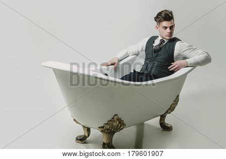 Handsome man or businessman with stylish haircut hair in fashion business wear vest tie shirt and pants relaxing in classic bath white tub on grey background
