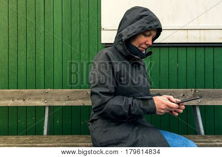 Authentic woman in winter jacket using mobile phone to type sms message outdoors while sitting on the bench on a cold day
