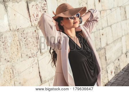 Young stylish woman wearing neutral blazer, hat and sunglasses walking on the city street in spring. Casual fashion, elegant look. Plus size model.