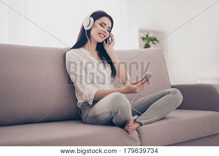 Portrait Of Young Charming Smiling Woman Sitting On Sofa At Home Holding Cellphone And Listening To