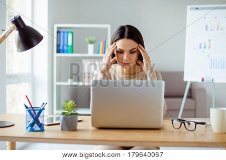 Thoughtful Pretty Tired Woman Sitting At The Table Touching Her Temlpes And Looking At Screem Of The