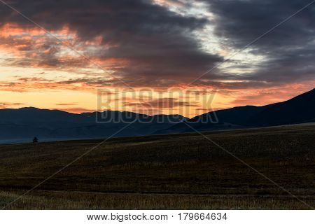 A picturesque landscape with mountains trees steppe and beautiful storm clouds with orange backlighting at sunset